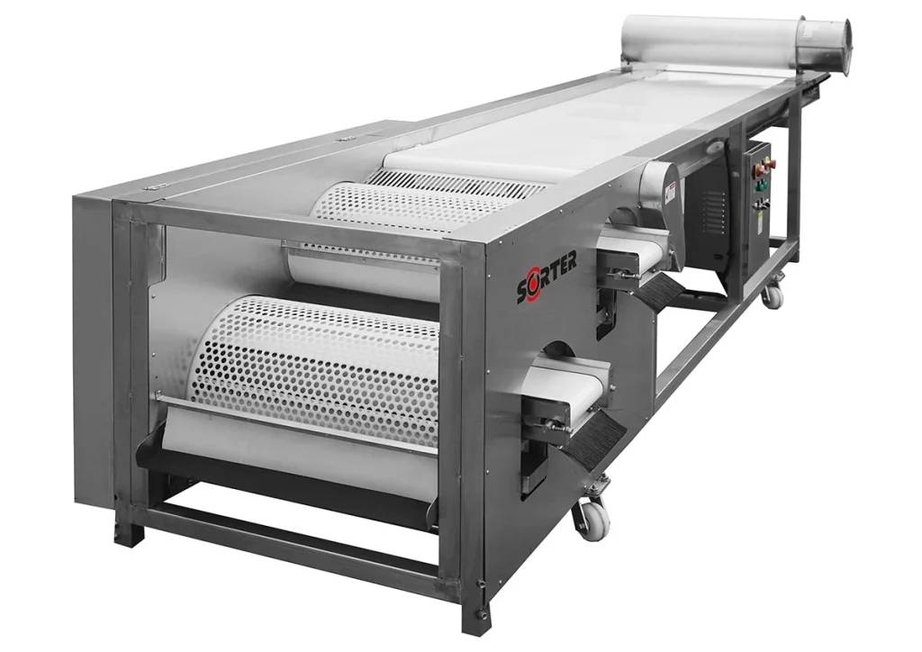 Duża przemysłowa maszyna do przetwarzania żywności z metalową ramą, dwoma perforowanymi bębnami obrotowymi, panelem sterowania i przenośnikiem taśmowym biegnącym przez środek. Maszyna jest na kółkach zapewniających mobilność.