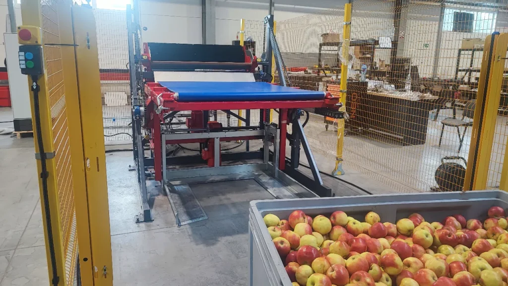 Pojemnik wypełniony jabłkami znajduje się w pobliżu przenośnika w zakładzie przemysłowym. Przenośnik ma niebieską taśmę, a obszar otaczają żółte bariery bezpieczeństwa. W tle widoczne są regały i sprzęt.