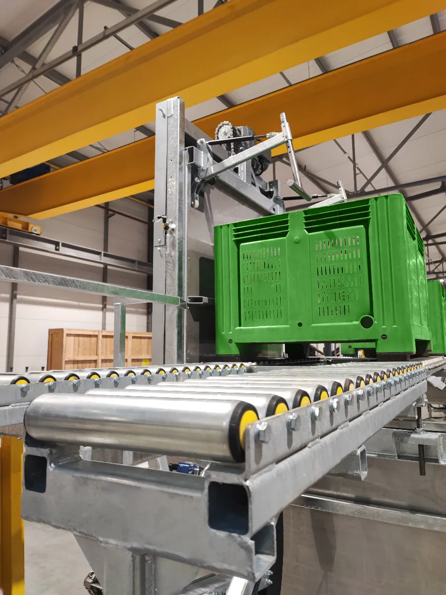 A green plastic crate moves along a metal roller conveyor inside an industrial warehouse with yellow support beams and a high ceiling.