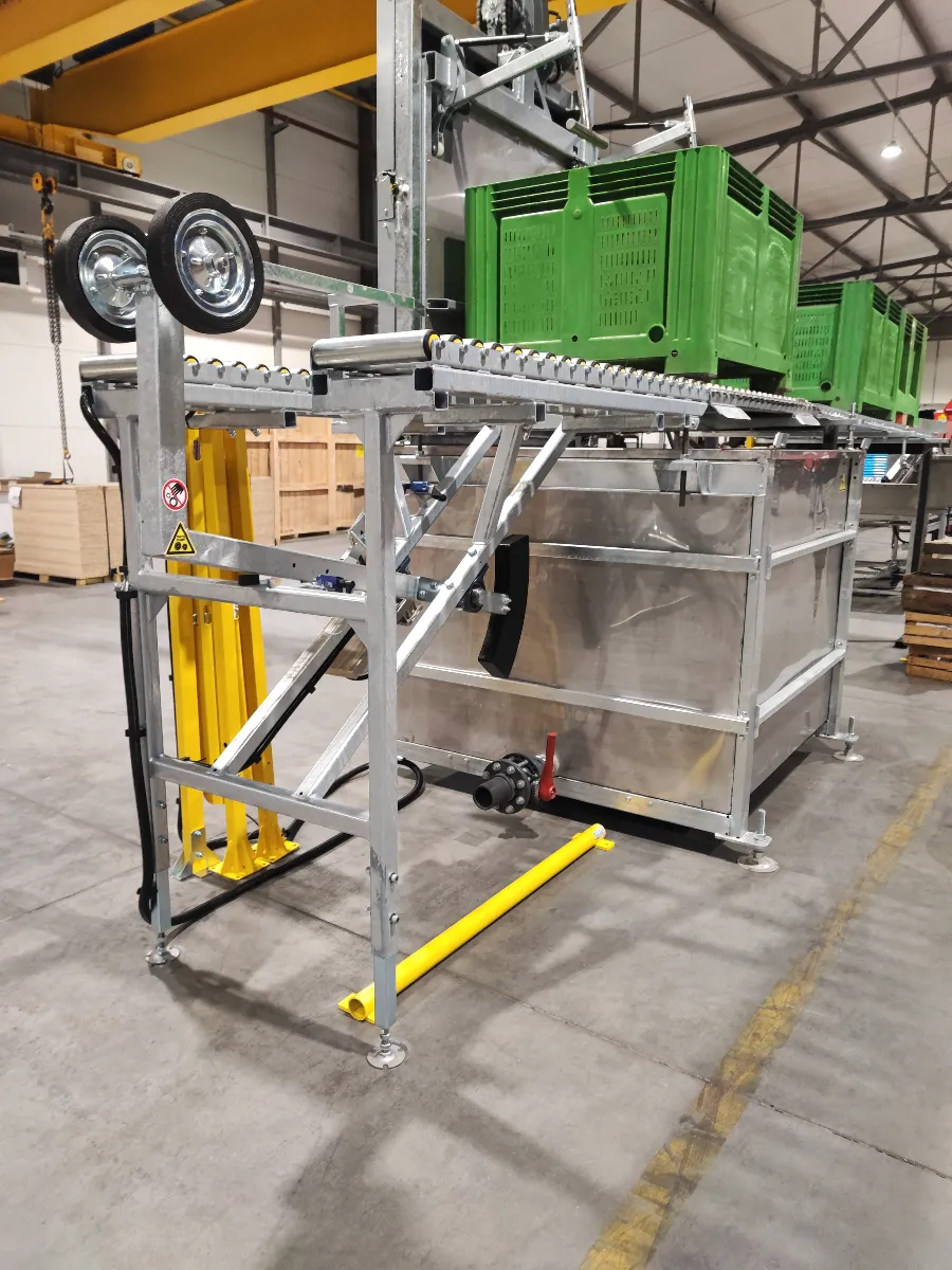 Industrial machine with metal frame, conveyor rollers, green crates on top, yellow upright support, wheels and warning sign, located in a warehouse with crates and pallets in the background.